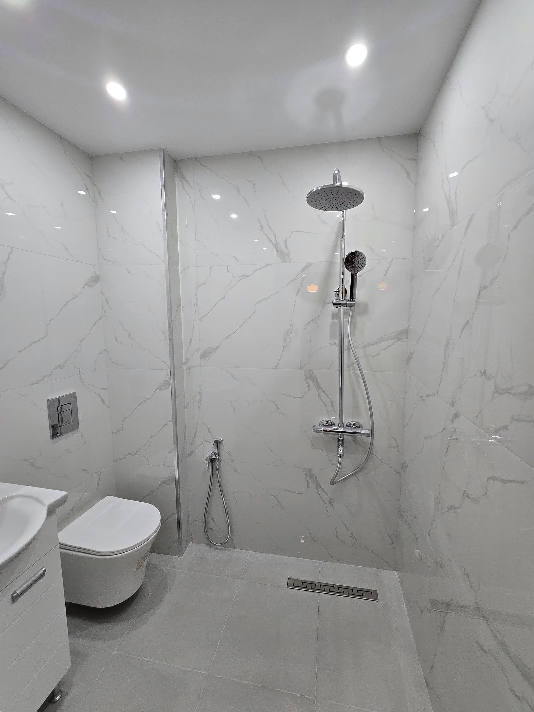 A bathroom with white marble-effect tiles: a walk-in shower with a linear drain, a wall-hung toilet and a bidet shower