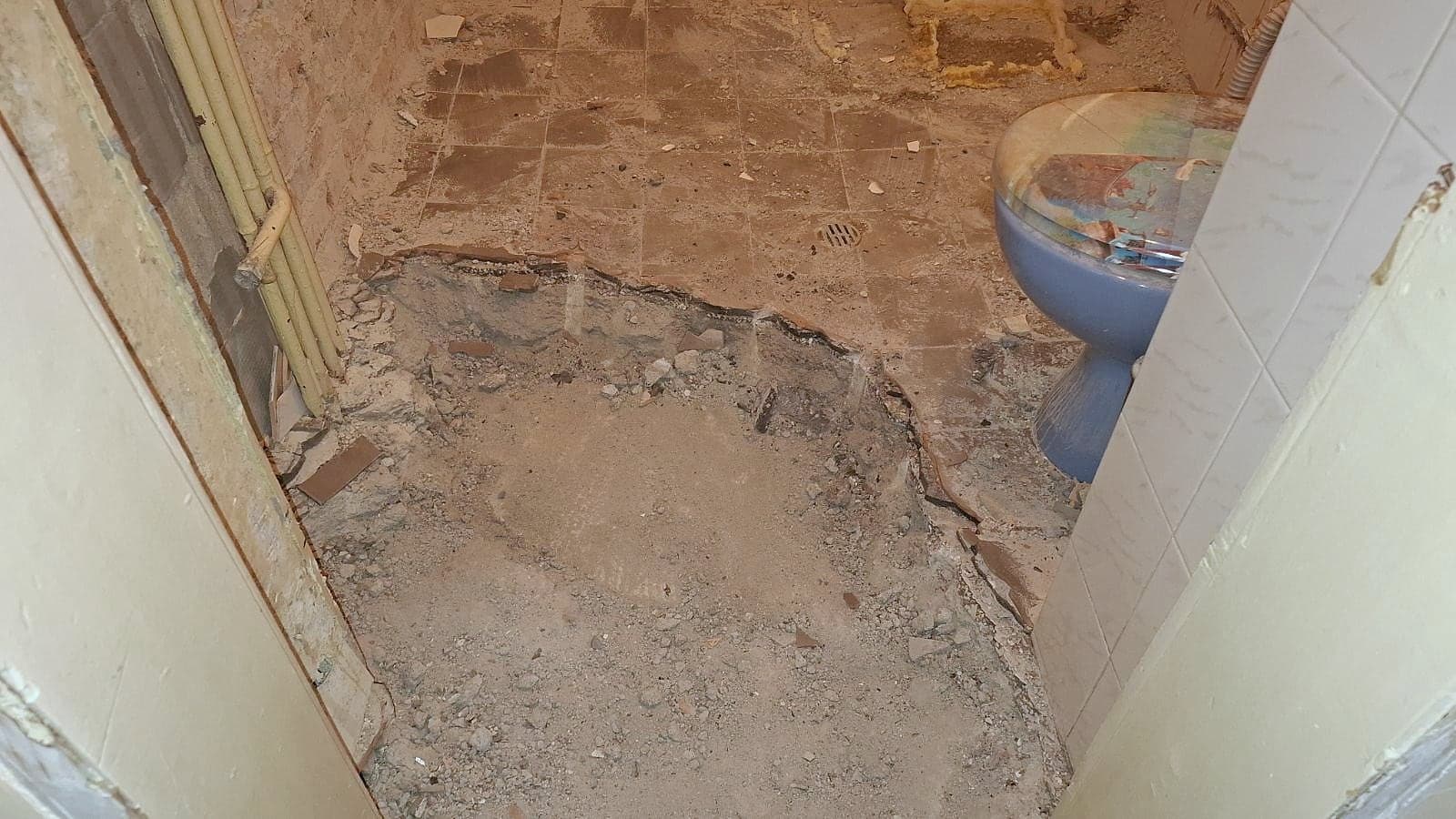 The broken-up floor of an old bathroom, with the screed chipped out, pipes along the wall and an old blue toilet