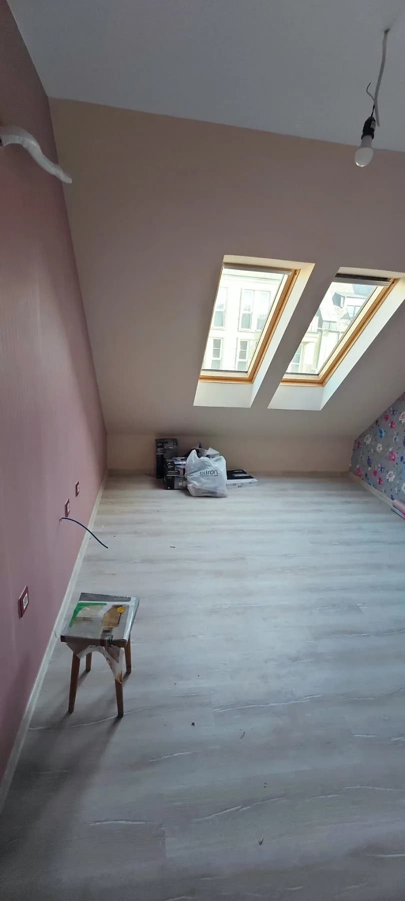 The same attic room from the other corner: two roof windows in the sloping ceiling, a pink wall on the left and the floral wallpaper at the far end on the right